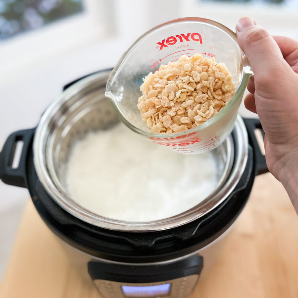 A measuring cup with rice crispy treats cereal
being poured into the marshmallow fluff in the Instant Pot